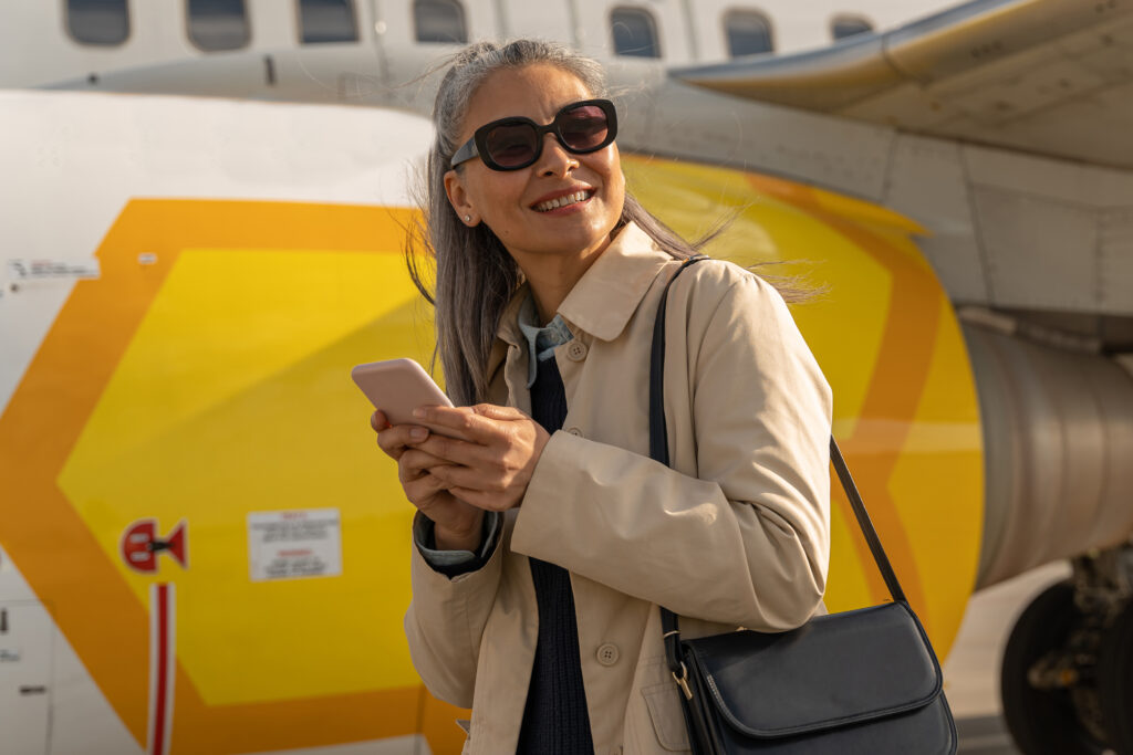 Joyful female traveler holding phone and smiling while standing at airfield near plane