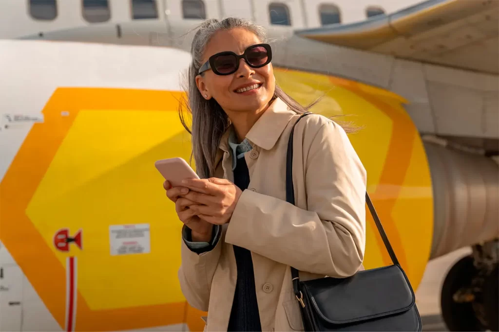 Joyful female traveler holding phone and smiling while standing at airfield near plane
